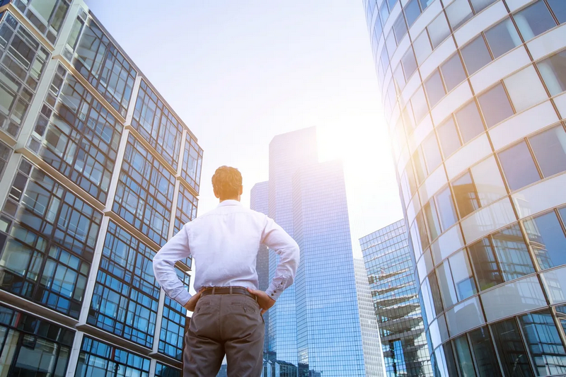 A man in a white shirt stands with hands on hips, looking up at modern skyscrapers in a city.