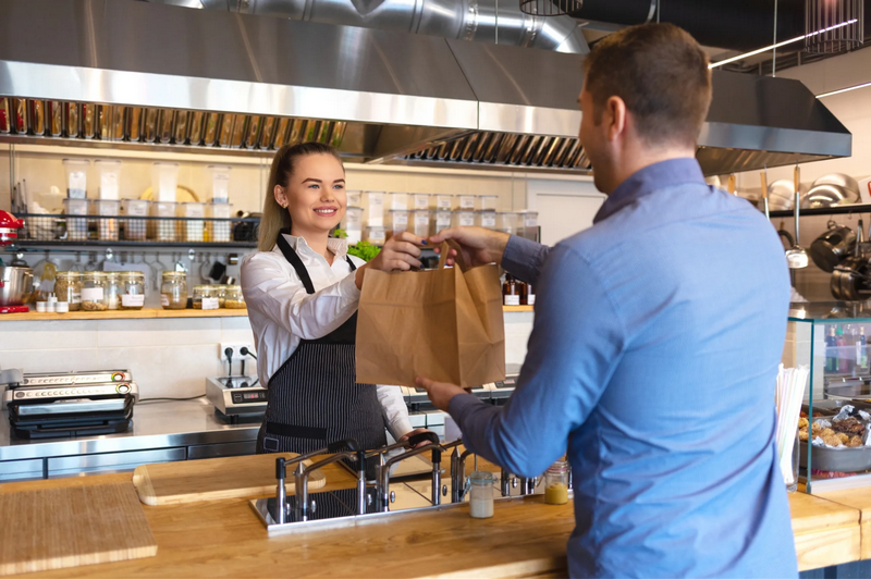 A cashier hands a takeout order to a customer in a modern restaurant kitchen.